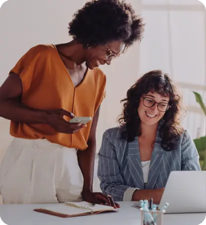 Two adults are smiling and working together at a desk with a laptop, notebook, and pens in a bright, modern setting.

Description generated by AI