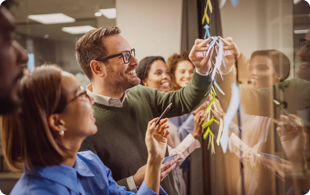 People in casual clothing are smiling and collaborating while placing colorful sticky notes on a glass wall in a modern office setting.

Description generated by AI