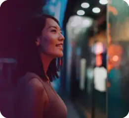A person with shoulder-length hair wearing a sleeveless top is smiling and looking at a brightly lit storefront or display window at night.

Description generated by AI