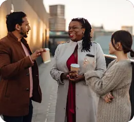 Three adults dressed in coats are standing outdoors in an urban setting, holding coffee cups and engaging in a lively conversation.

Description generated by AI