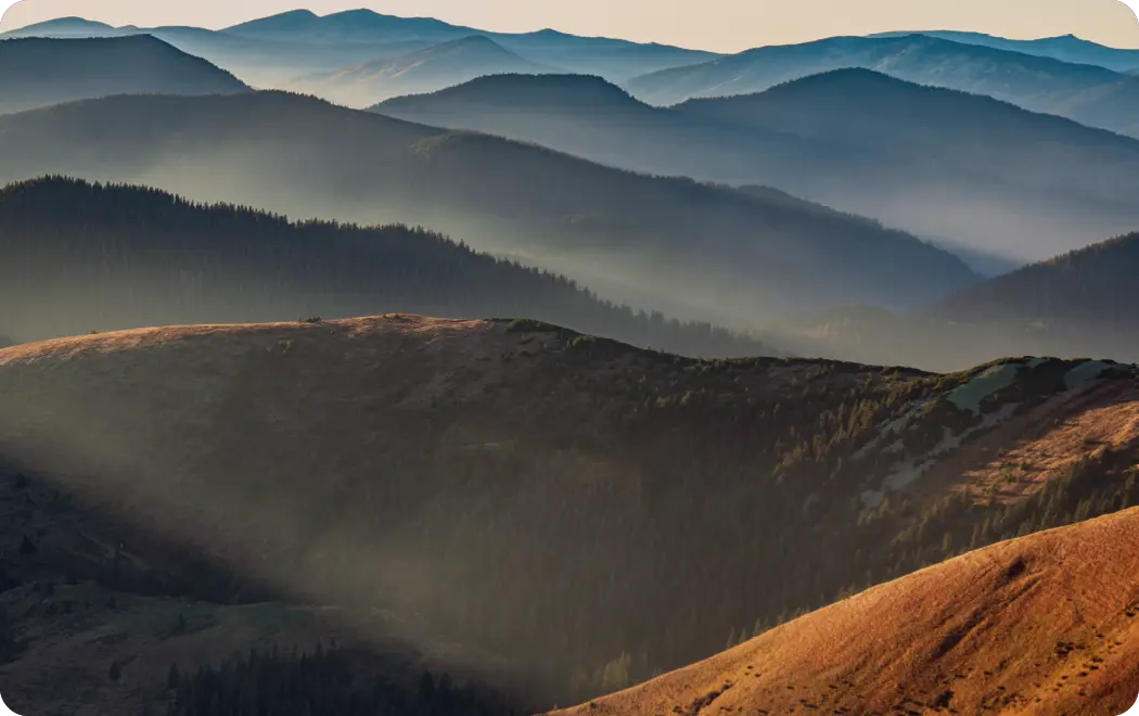 Layers of misty blue and green mountain ridges stretch into the distance under soft sunlight.

Description generated by AI