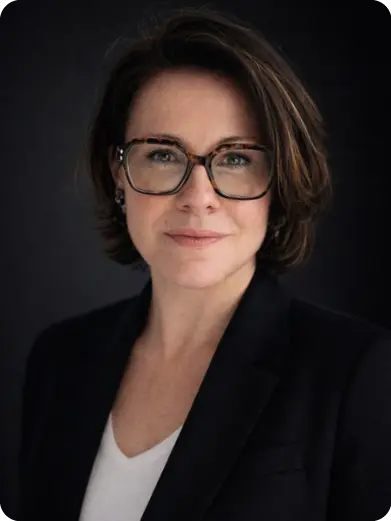 Person with short hair and glasses wearing a black blazer and white shirt against a dark background looking calmly at the camera.

Description generated by AI