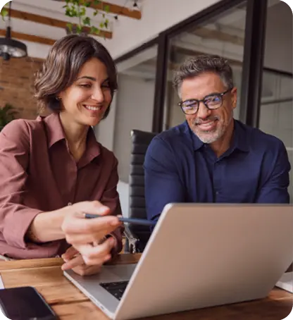 Two adults are smiling and looking at a laptop while one points at the screen with a pen in a modern office setting.

Description generated by AI