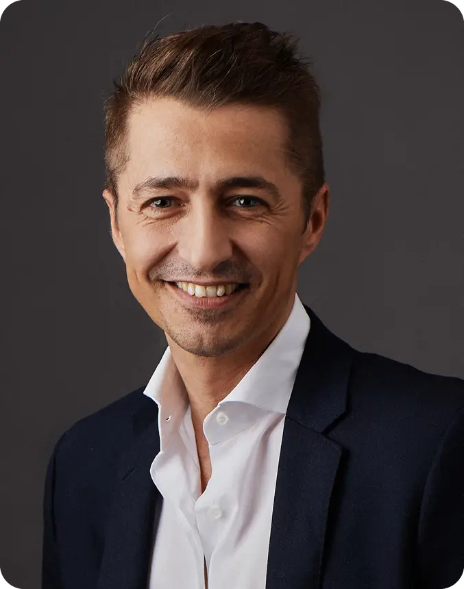 A person with short styled hair is smiling and wearing a dark blazer over a white button-up shirt against a dark background.

Description generated by AI